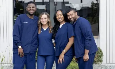 Wide-angle group photo of Herzing University medical assisting students smiling in navy scrubs outside a campus building, emphasizing teamwork and inclusivity.